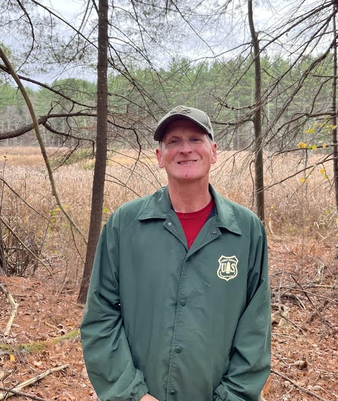 A smiling man wearing a scouting cap and a US Forest Service jacket stands in a field. There are a few bare trees right behind him and a line of conifers in the background.