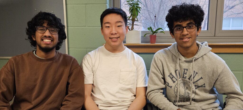 Three smiling young men sit in front of a window. A corkboard in the background indicates that they are probably at ABRHS.