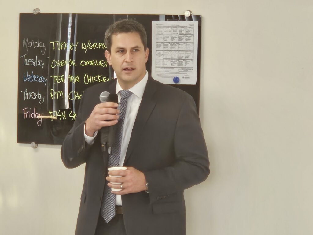 A man in a suit and tie holds a mic in one hand and a cup of coffee in the other.