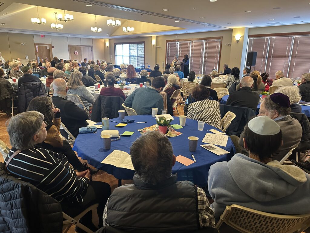 The Beth Elohim meeting room is set with several round tables, all of which are full of people listening to the speaker.