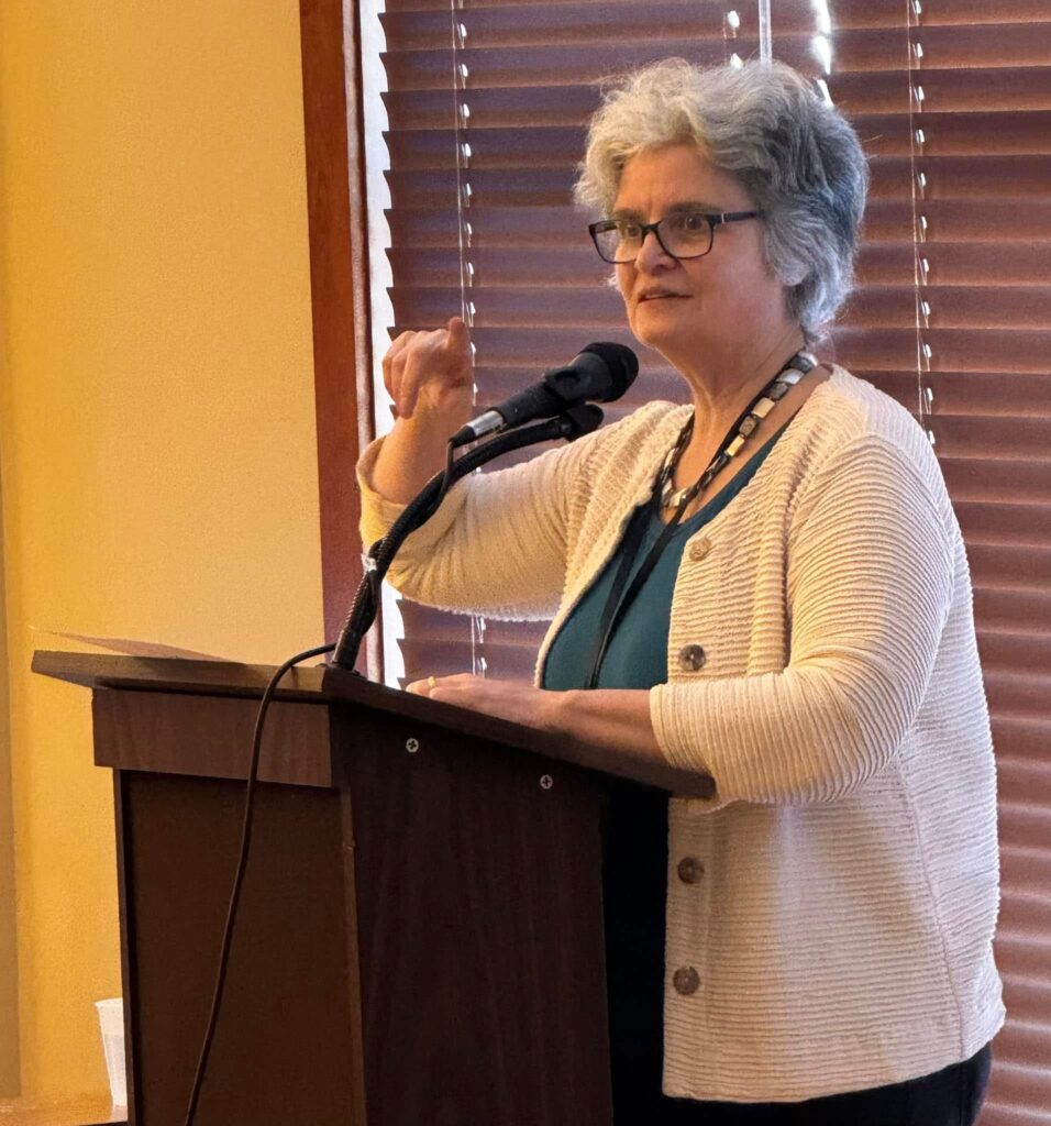 A woman wearing a white sweater and green shirt speaks at a podium.