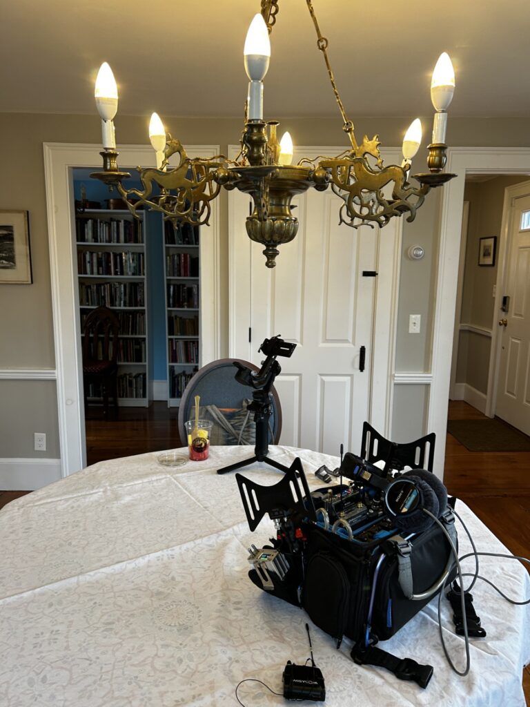 A bag of camera equimpent sites on a dining room table. A chandalier is above the table and you can see a library in the background.