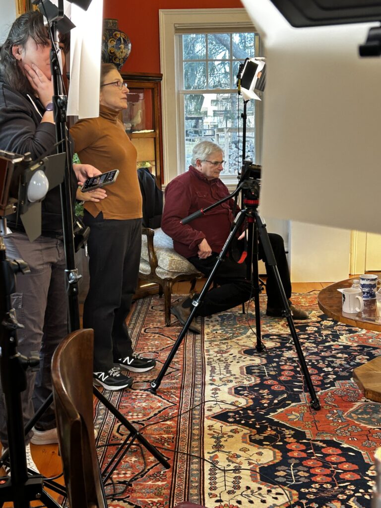 A woman in a brown turtleneck stands looking at a piece of equipment. Behind her, a man sits on a living room chair. In the foreground, anotherperson stands, mostly hidden by more equipment.