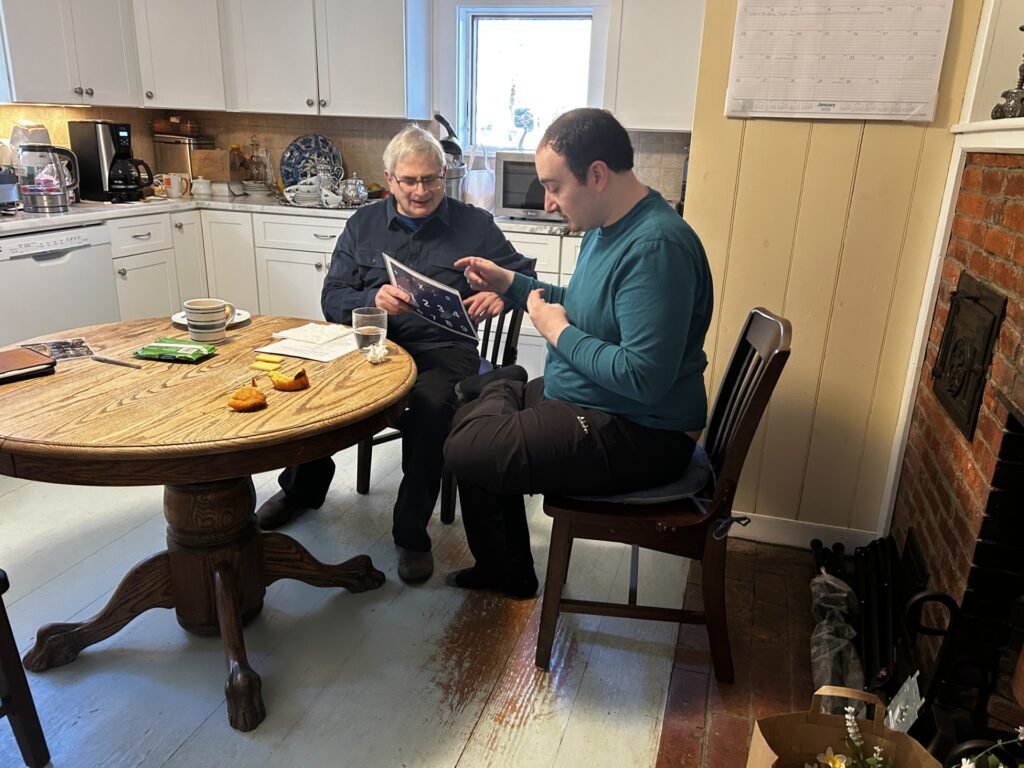 Two men sit at a kitchen table. An older man on the left is holding a letter board. The younger man is pointing to letters on the board.