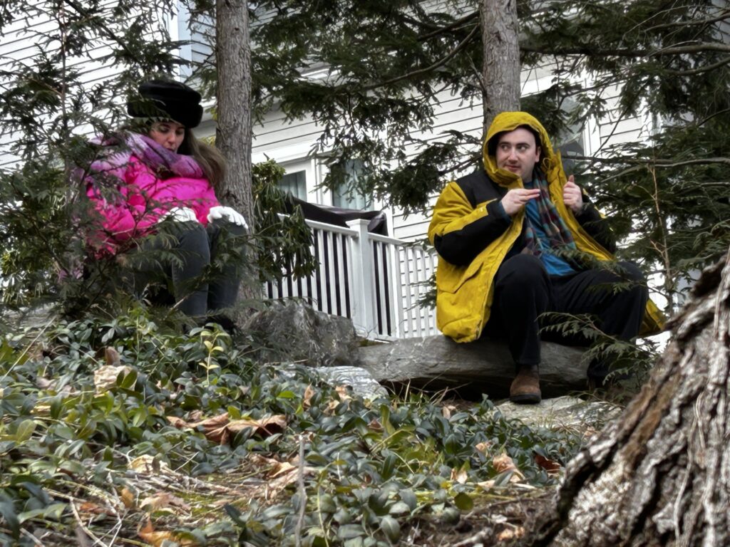 A woman in a pink parka sits under a tree. To her right, a man in a yellow parka sits on a rock and looks at her sidewise.