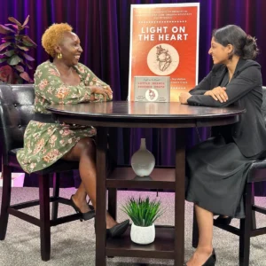 Two women sit at a bable and talk. A Light on Heart poster sits between them.