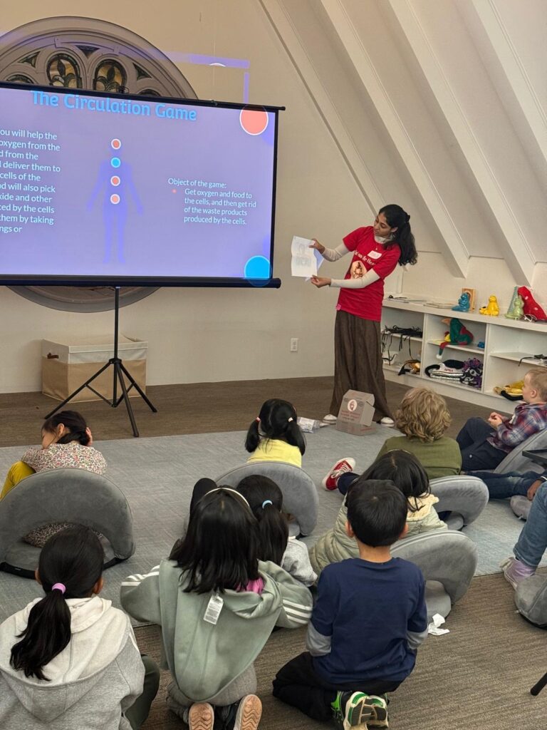 A young woman wearing a red t-shirt stands in front of a screen with text on it. A bunch of kids are sitting on the ground listening.