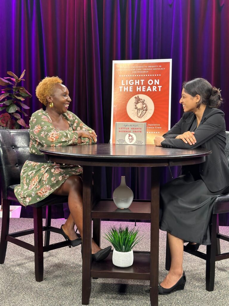 Two women sit at a bable and talk. A Light on Heart poster sits between them.