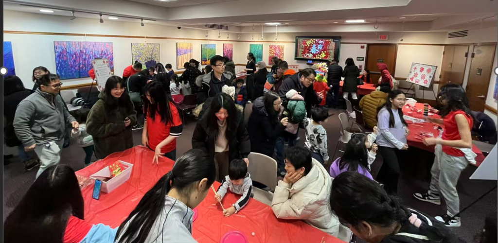 A busy scene with lots of people walking around and sitting at tables with red tableclothes. The tables have art projects and games that people are using.