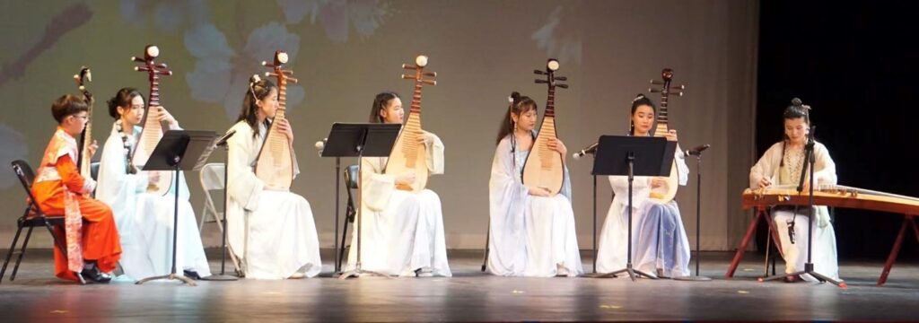 One boy wearing a red suit and five women in white dresses play the pipa - a lute-like instrument. On the right, another woman in whote plays a guzheng, which is a long stringed instrument on legs.