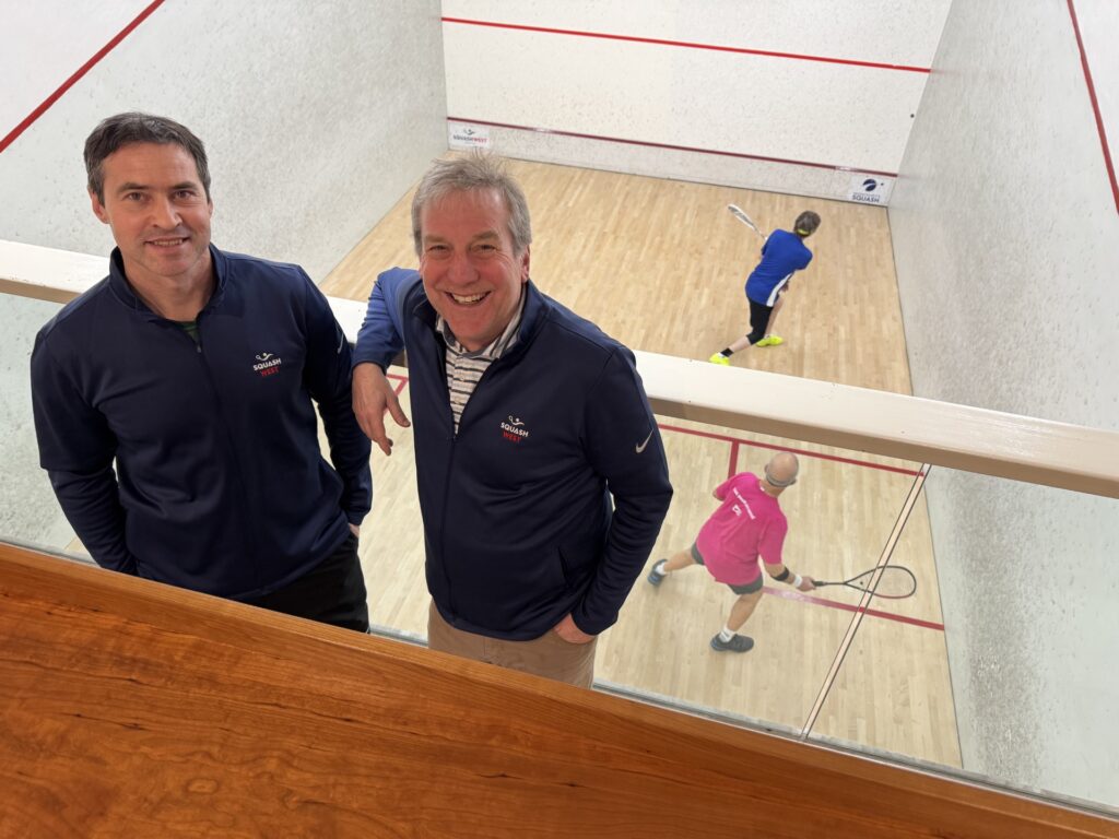 Two men are in the balcony above a squash court. Behind them, a couple of people are playing squash.