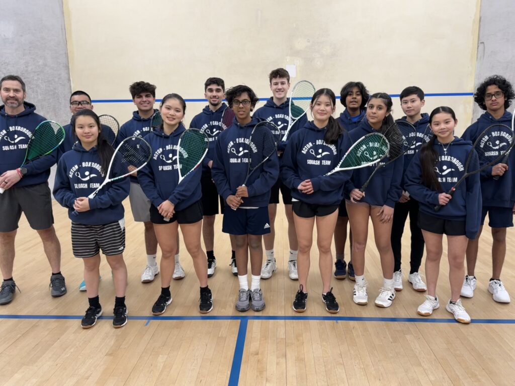 A group of kids, all holding squash rackets and wearing blue AB Squash Team sweatshirts, stand in a squash court with their coach.