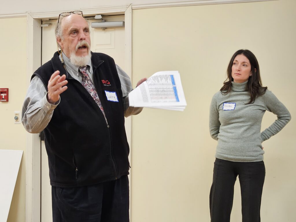 A tall bearded man in a black vest is holding a paper full of numbers and speaking. A woman in a gray sweater is standing off to the side.