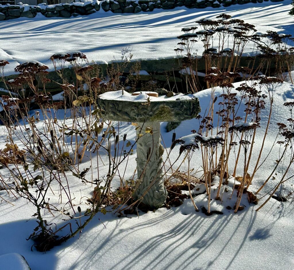 A stone birdbath in the snow. A stoone wall is behind it.