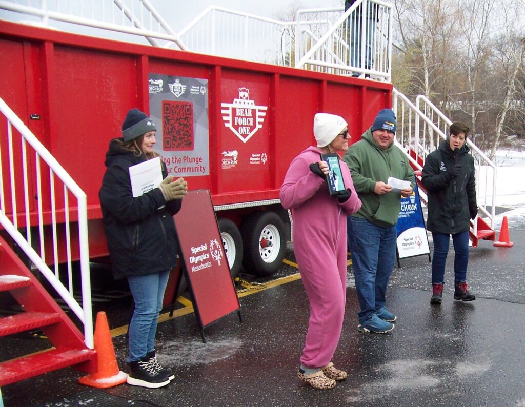 A woman wearing a pink onesie and holding an award stands in front of the plunge tank. Several other people are with her to present the award.