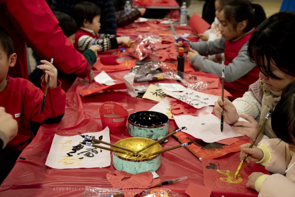 A bunch of young children at a red covered table. Some seem to be trying to write Chinese characters, a little girl would rather paint a gold kitty.