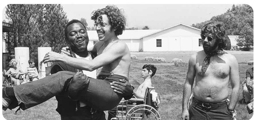 In the background, a large white building. In the foreground, a young man in a wheelchair, and one large smiling man carrying another happy man (possibly from the bus to his wheelchair). Most people in the picture are shirtless.