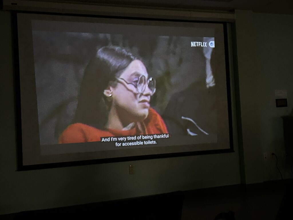 A woman with brown hair and large glasses. According to the closed captioning, she is saying " And I'm very tired of being thankful for accessible toilets."