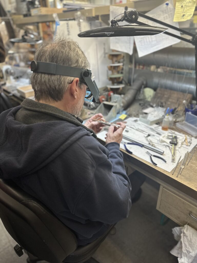 A man at a crowded workbench works on what appears to be a piccolo.