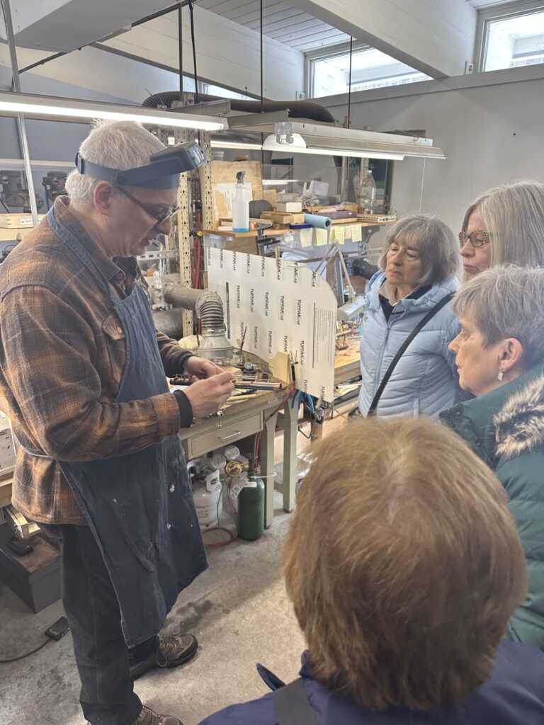 A man in a work apron shows a group of women a piece of a flute that he's working on.