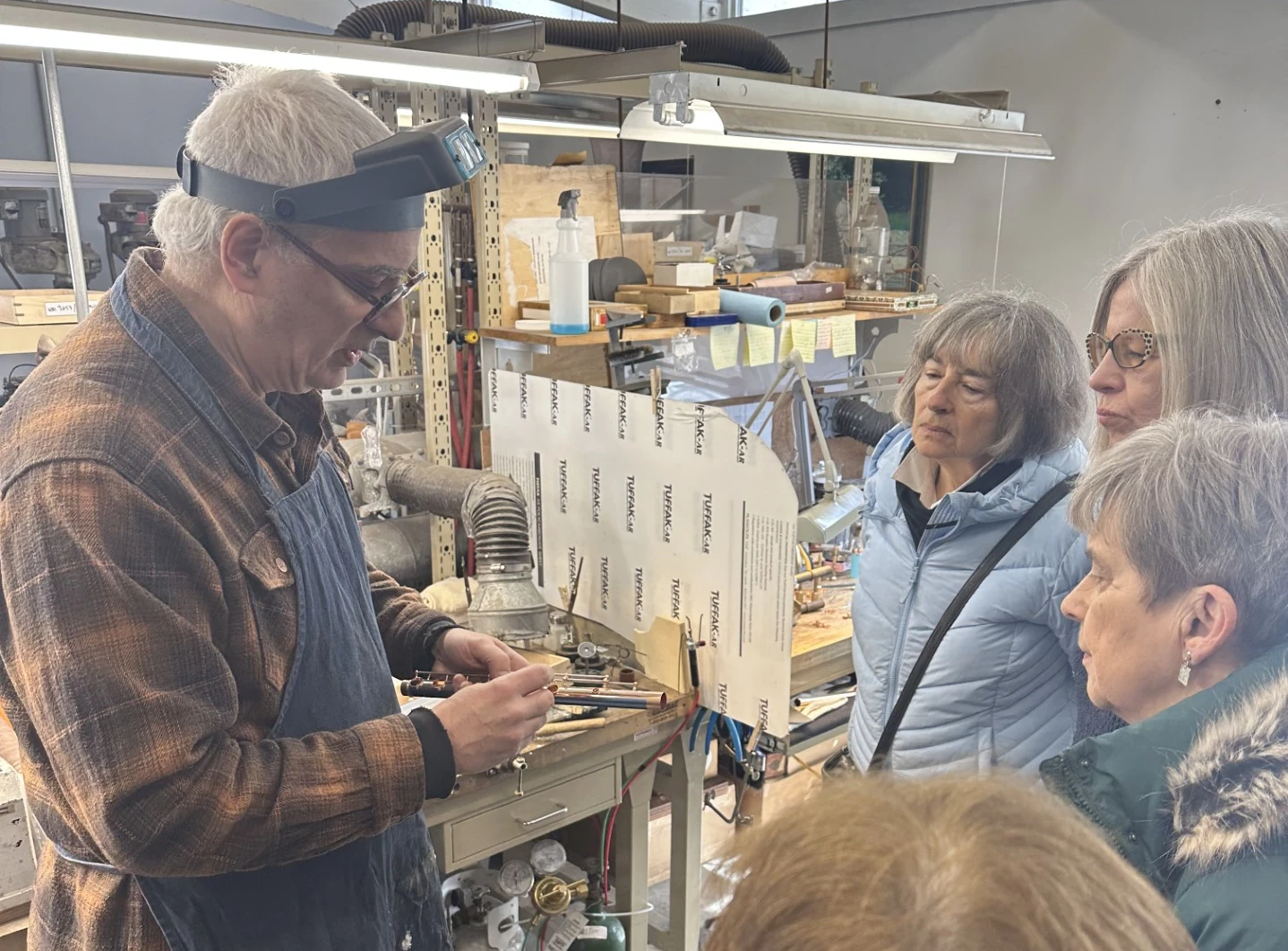 A man in a work apron shows a group of women a piece of a flute that he's working on.