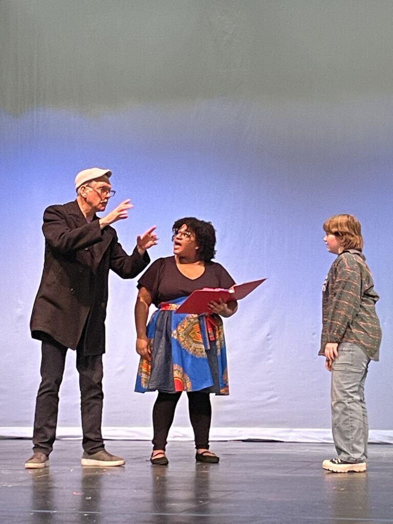 A white man in a cap and white mustache signs while his ASL Interpreter, a Black woman in a blue patterned skirt and a student look interested.