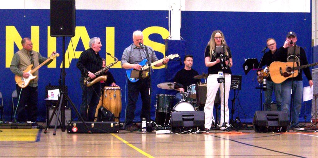 A band of older musicians plays against the yellow and blue AB backdrop.