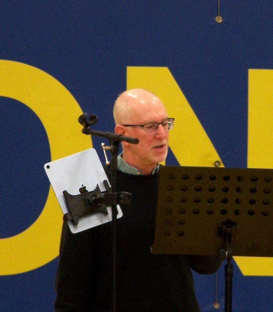 An older man with glasses speaks in front of a music stand.