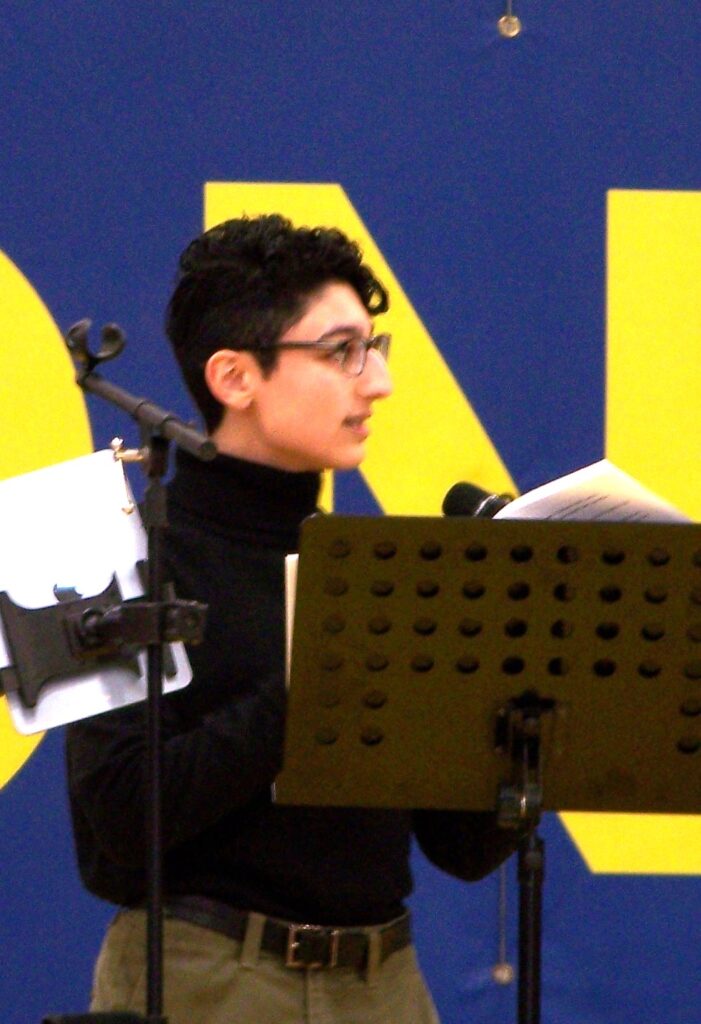 A young man with curly hair and glasses speaks in front of a music stand.